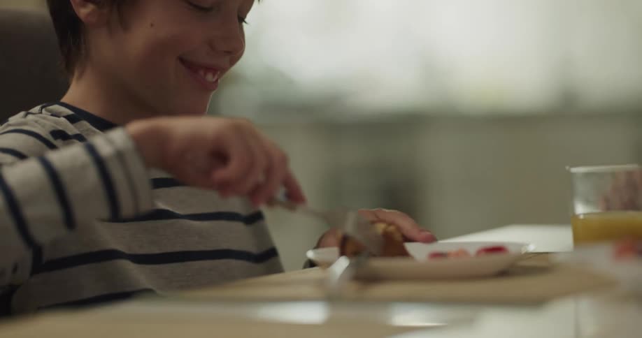 A young boy at home laughs and talks as he eats and then his mom reaches over and takes something off his plate