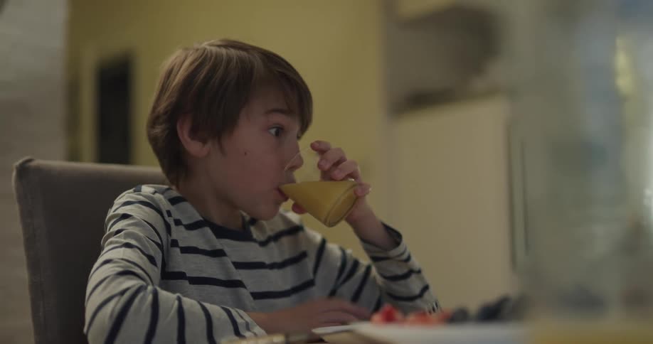 A young boy sits at home and talks as he eats and drinks during a meal