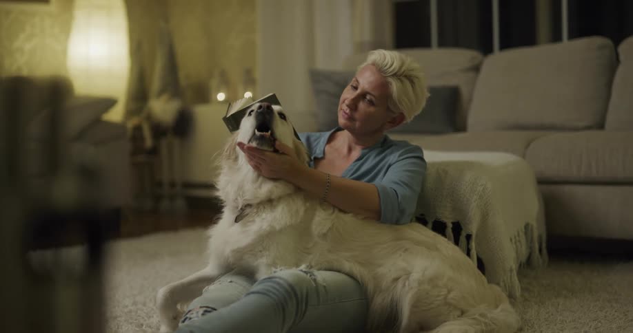 White woman sitting at the floor in her living room reading a book and petting her dog