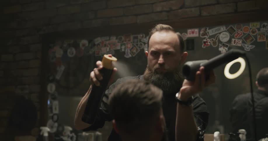 Male hair stylist sprays hairspray on a clients hair while holding a hair dryer in his other hand