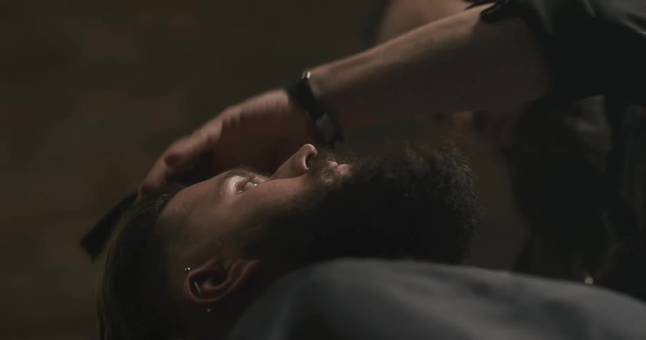 A barber trims the sides of a mans face as he leans back in a salon chair