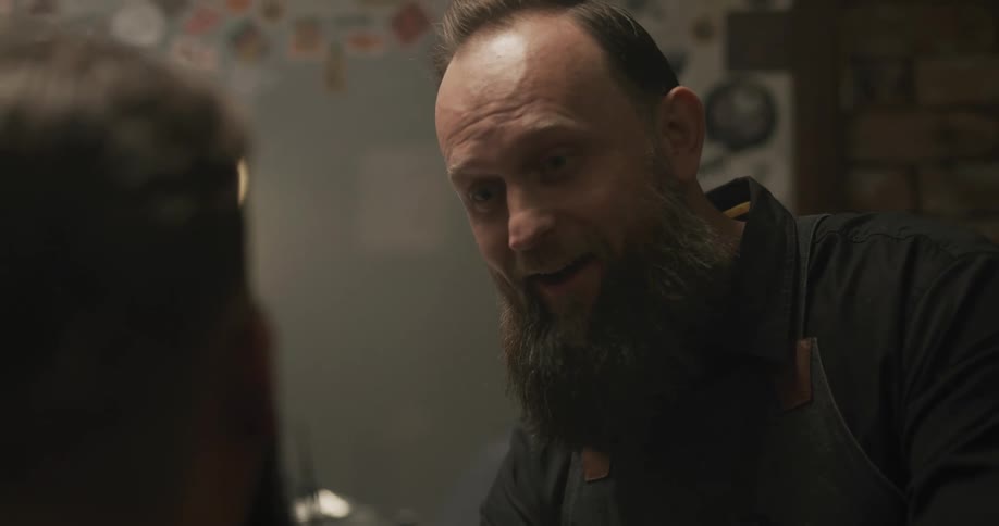 Barber with black shirt apron and beard talks to man sitting down in barbershop