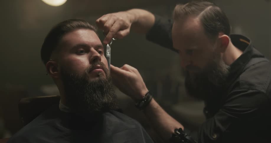 Barber trimming customers beard with long scissors in a barber shop
