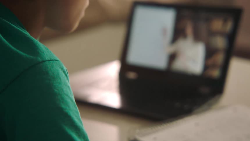 A young boy writes in a notebook sitting beside a laptop playing a video