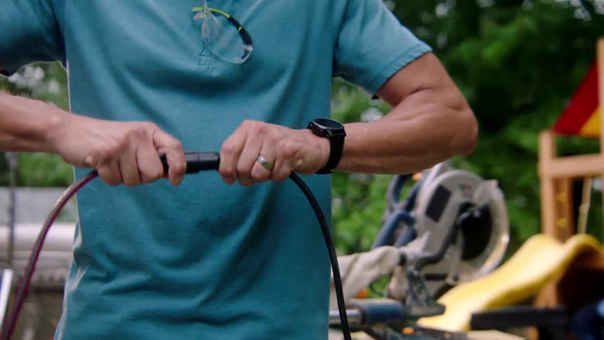Man connects an electrical cord to an extension cord while standing outdoors near an electric saw