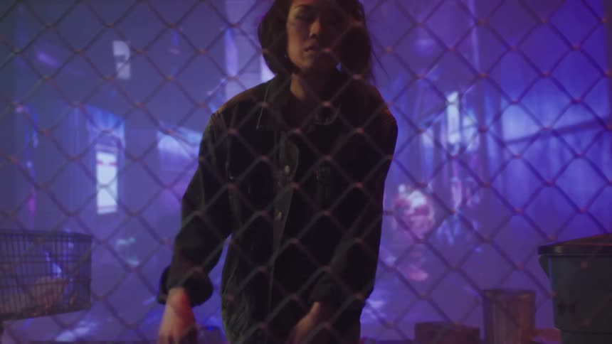 A woman with a jacket walking up to a fence and staring with garbage cans and a shopping cart in the background