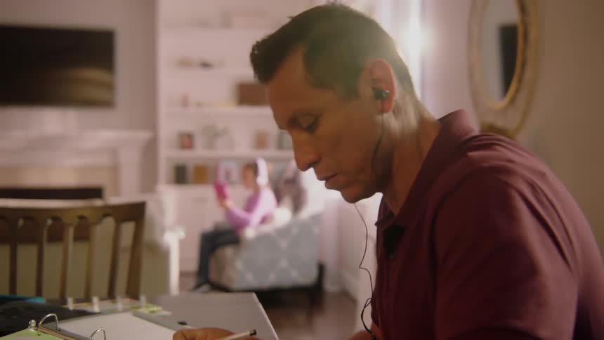 Father sitting at the table while studying and listening to music while his daughter is at the background listening to music and using her smartphone