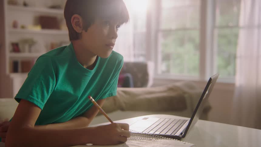 A young boy sits at home in front of a laptop and does school work