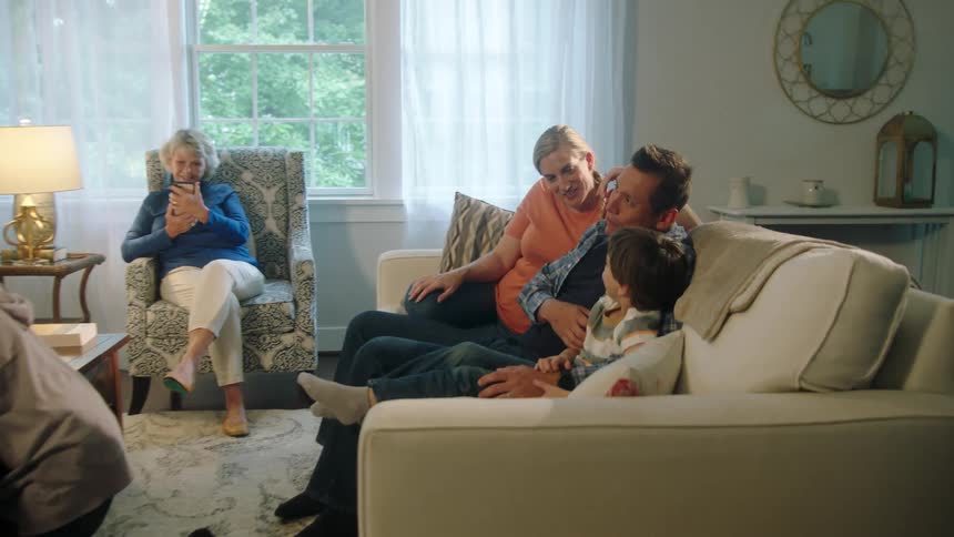 A mom and dad sit on a sofa with their son while their other children play a game and grandma sits in a chair talking on her phone