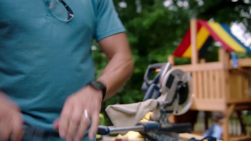 A man with a wristwatch plugging in an electric saw with children and a swing set in the background