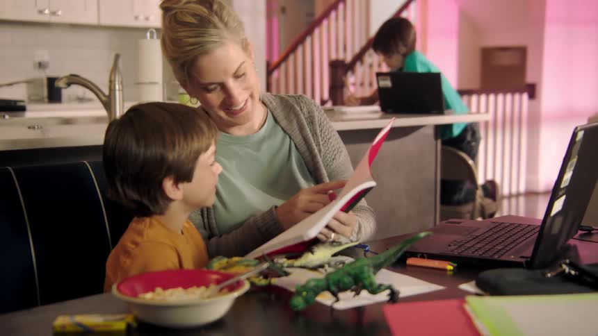 A woman points in a red notebook as she helps her son with homework as the family sits in the kitchen
