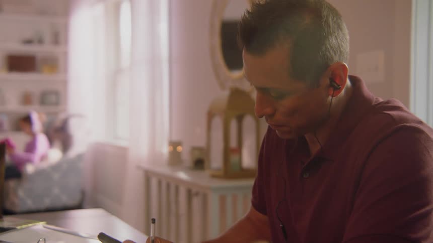 A man sitting at table in the living room of a houses writes on a notepad while looking at the screen of his smartphone before rising from the table and walking away