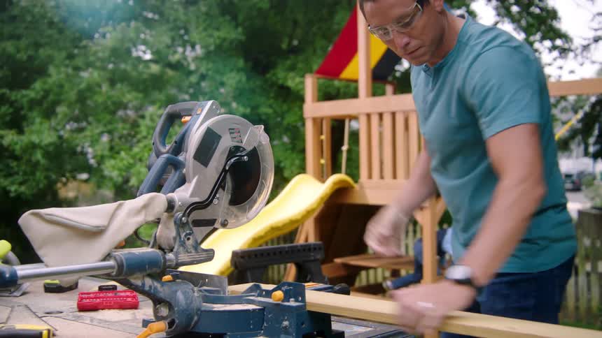 A man cuts a piece of wood in his backyard as his children play on a swing set