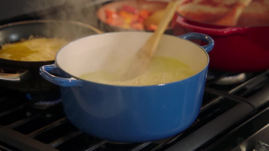 A hand uses a slotted spoon to stir a pot with pasta in water on a gas stove