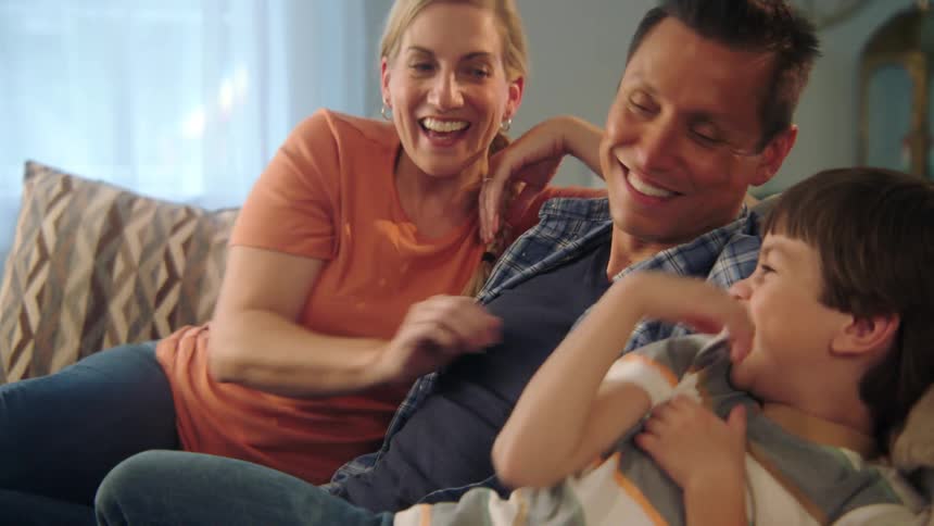 A man a woman and a boy sitting on a couch with a woman tickling the boy with a window in the background