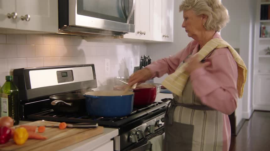 An elderly woman stirs two pots of food on her stove as she cooks and then wipes her hands on a towel