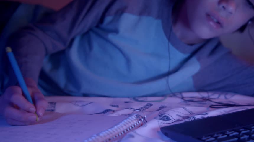 A boy in a bedroom laying on a bed looking at a laptop and writing on a notebook