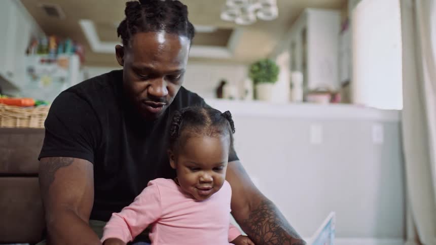 Black man sits on the floor with his young daughter and reads her a book