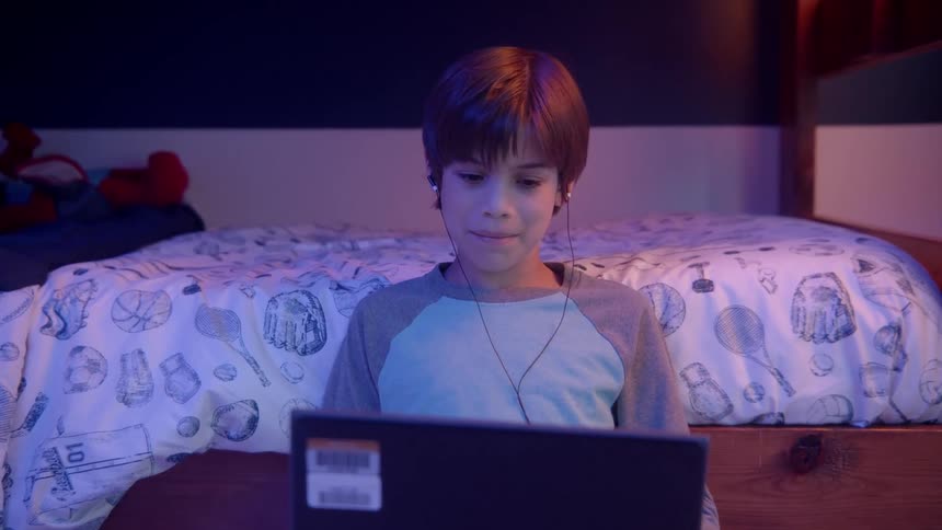A boy wearing earbuds talking to someone on a laptop while sitting in the bedroom floor