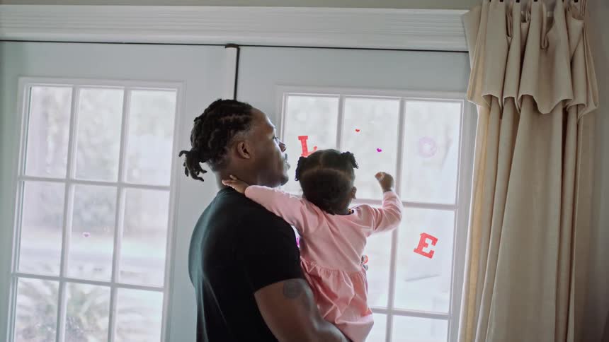 A little girl reaches for a sticker on a window while being held by a man standing indoors during the day