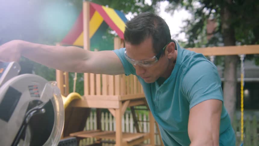 A man cuts wood with a circular saw while children play on a swing set and slide behind him