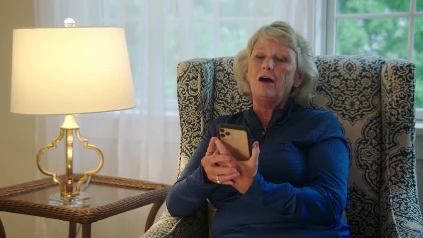 An older woman sits in an arm chair having a video call on a cell phone