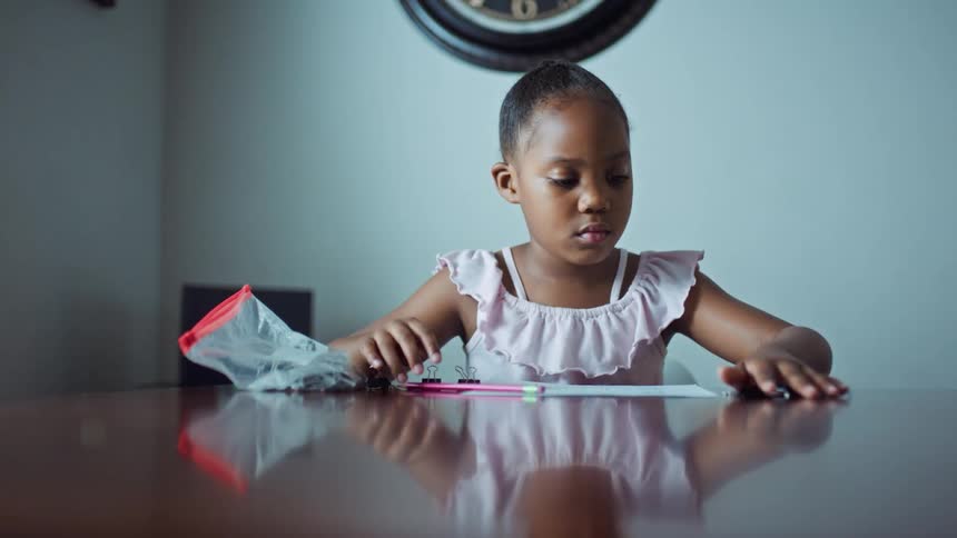 A girl in a room sitting on a chair at a table with a clock and a wall in the background