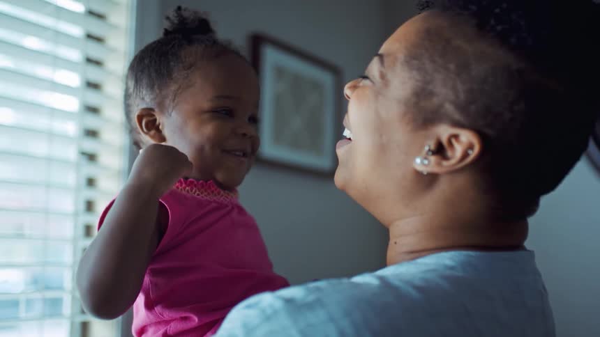 A woman holding a little girl in a room with a window a clock and a wall in the background