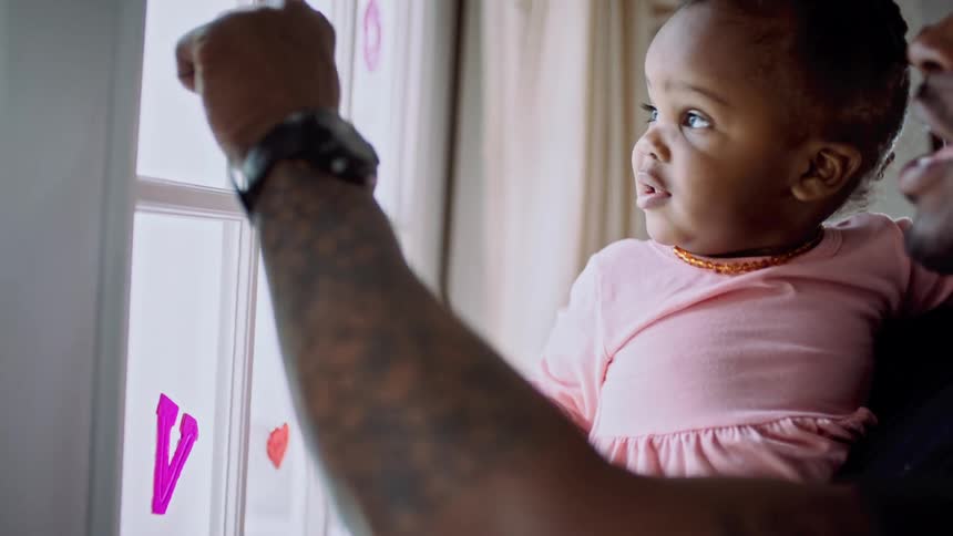 A man holds his toddler daughter and shows her valentines decorations on their window