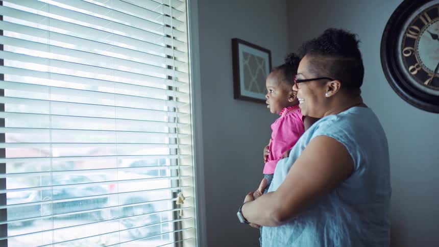 Mother holding her daughter next to a big window while the baby is waving and the mother laughing and talking