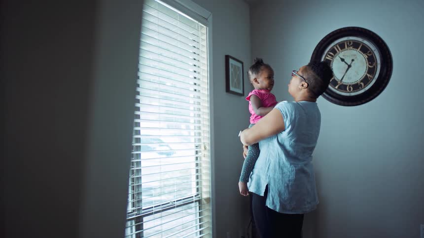 A woman in a room holding a little girl next to a window with a wall and a clock in the background