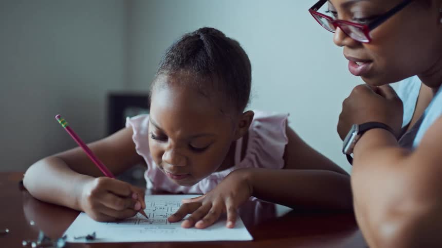 A woman with glasses sitting at a table teaching a little girl who is writing on a paper with a pencil