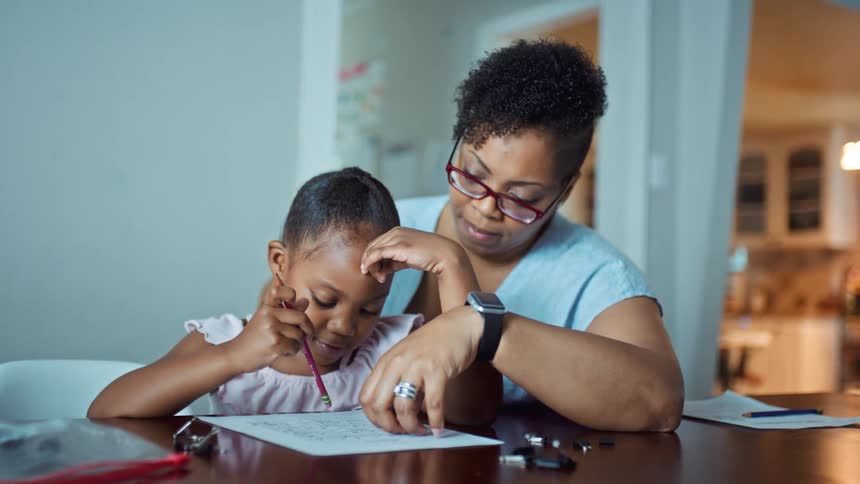 A woman explains an educational worksheet to her daughter as they sit together at a table in a house