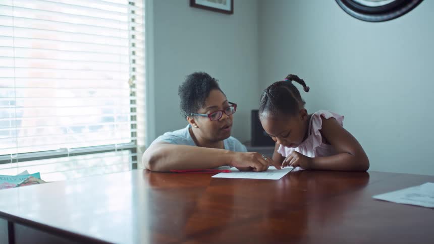 A mom kneeling next to her daughters helping them read and learn at the dining room table
