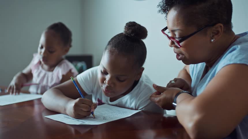 A woman sitting at a table with her daughters speaks to them as they fill out educational worksheets