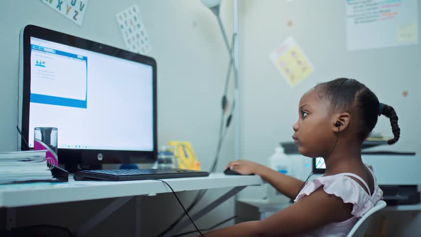A young girl sits at a desk and looks at a computer as she does school work