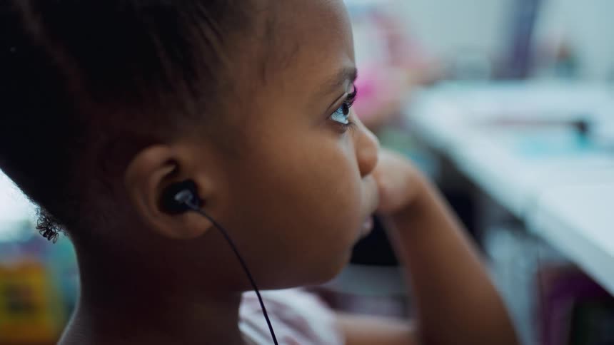 A young girl sits with her earbuds in and her head resting on her hand as she looks straight ahead