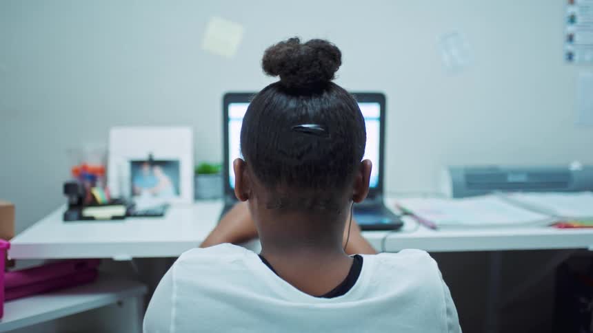 A girl sits at a desk facing a wall with an earbud in as she looks at her laptop