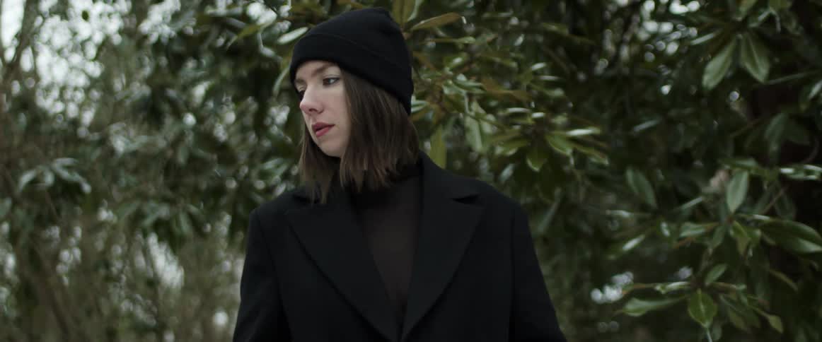 Woman wearing a black knit hat and a black coat is looking around with many trees behind her