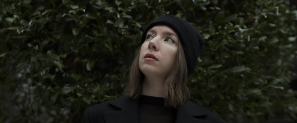 Woman wearing black knit hat is standing among a lot of trees and looking around