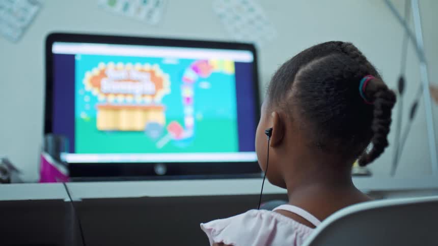 A young girl wears earbuds while playing a colorful video game at her desk