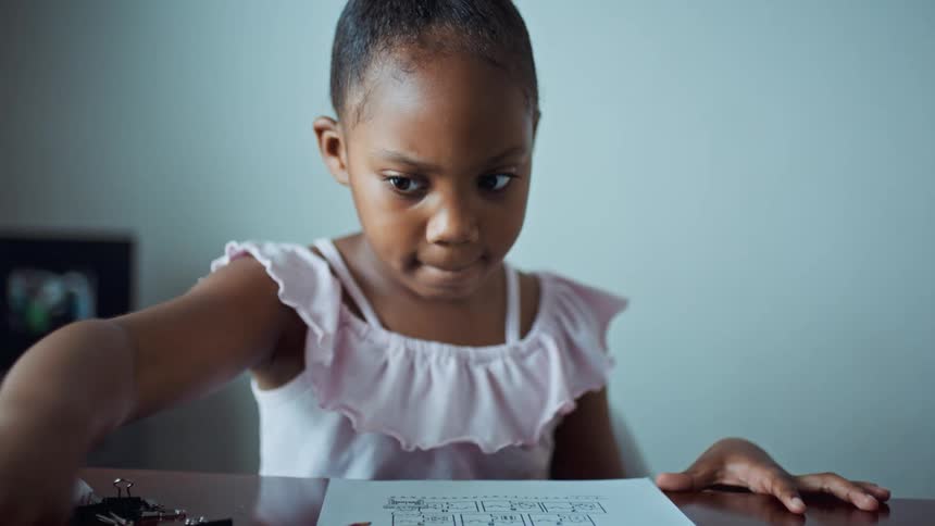 A girl in a room sitting on a chair at a table looking at a paper with a wall in the background