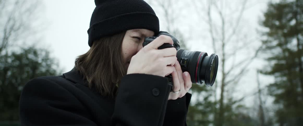 A woman with a hat and jacket and camera taking pictures with trees the sky and clouds in the background