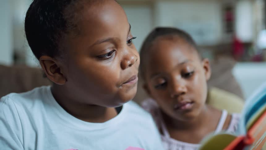 Two girls in a room sitting on a couch and reading a book