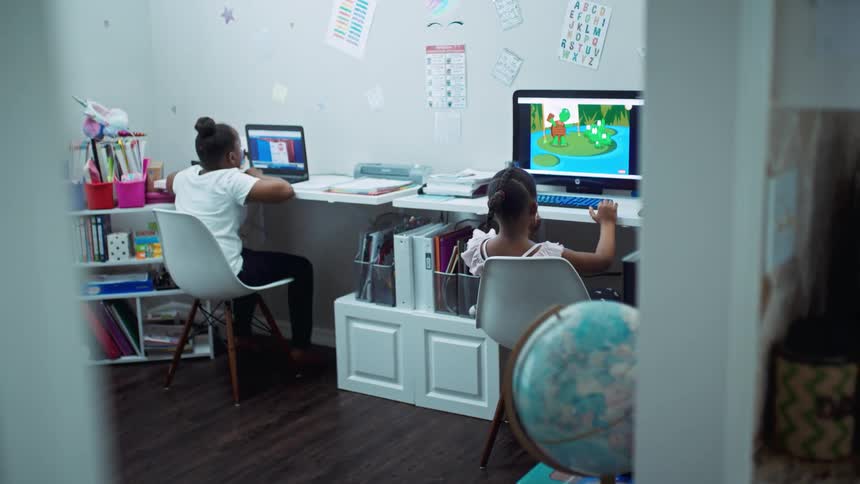 Two girls in a room sitting on chairs next to desk with one using a computer and another one using a laptop