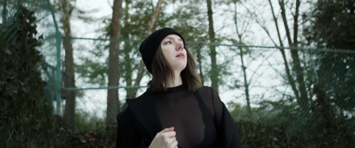 A woman wearing a sheer black blouse under her coat looks around at trees as she walks