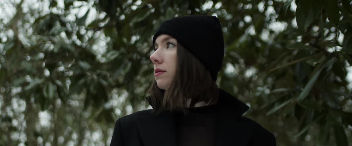 A woman wearing a black hat and black clothes looks up at the green leaves and trees as she walks