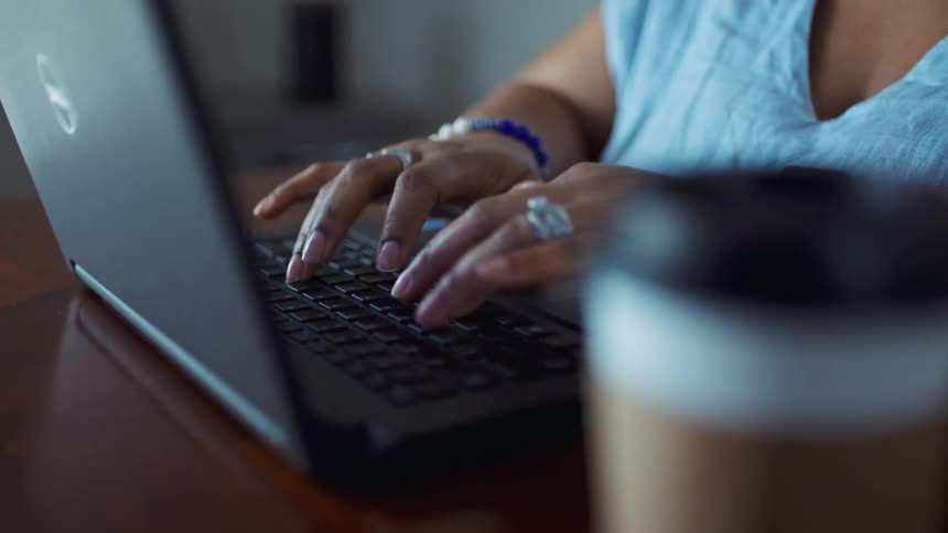 A woman types on a laptop keyboard with a coffee cup on the table