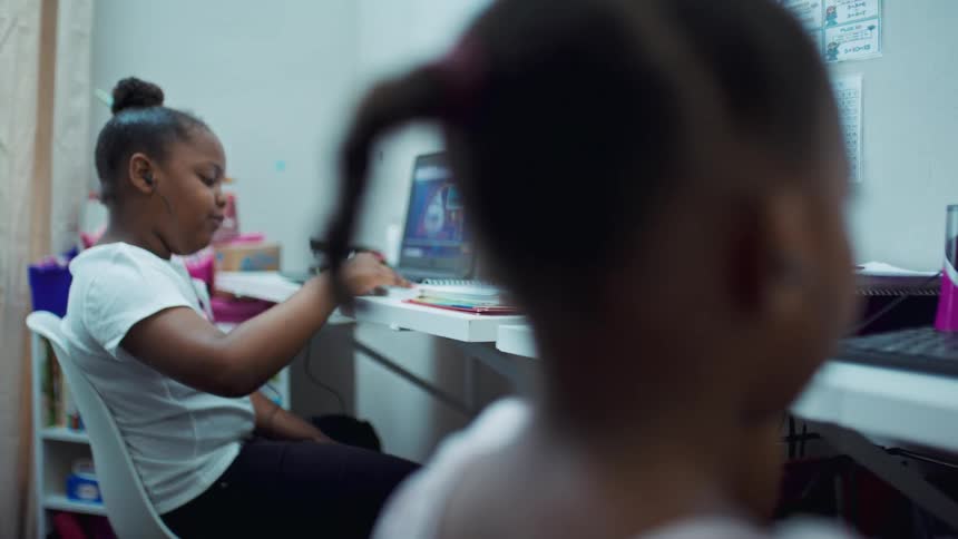 Two girls in a room sitting at desk one in front of a computer and the other one in front of a laptop