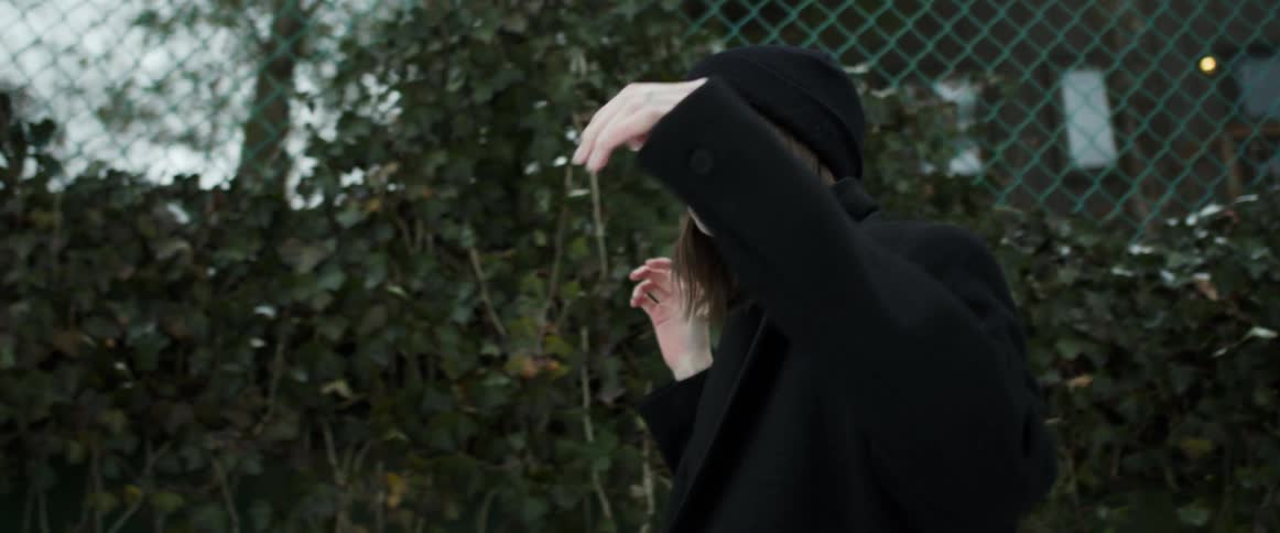 A woman with a jacket and a hat walking next to a fence with trees the sky and clouds in the background